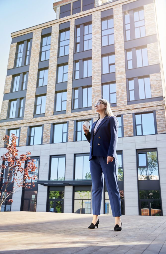Ambitious mature woman in styish outfit and high heels posing near tall office building